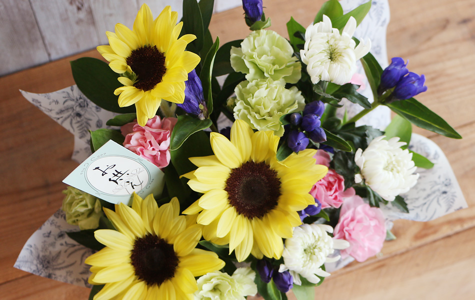 明るい黄色だけでなく、お供え花の定番色である紫（リンドウ）や白（菊）、優しいピンクをバランスよく配置し、暗くなりすぎず、ご遺族の心も和ませるような落ち着いた華やかさがあるデザイン